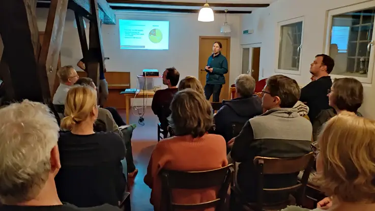 Vor einer Menschengruppe in einem Seminarraum zeigt ein Mann mit Pferdeschwanz eine Beamer-Präsentation.