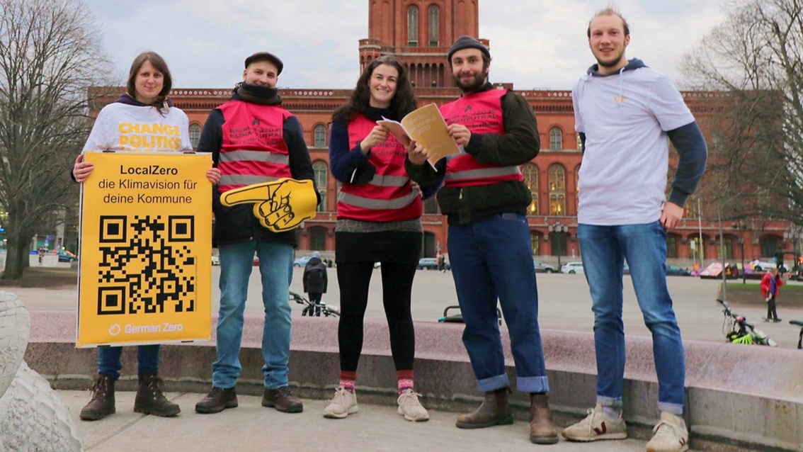 Fünf junge Menschen stehen vor einem wilhelmnischen Backsteingebäude auf einem Platz, eine der beiden Frauen hält ein Plakat mit der Aufschrift  «LocalZero – die Klimavision für deine Kommune» und einem QR-Code in den Händen