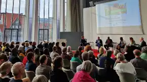 In einem hohen Saal mit großen Fenster lauscht eine große Zuhörerschar zwei Menschen auf einer Bühne, hinter diesen eine Präsentation.