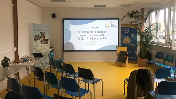 Ein großer, freundlich gestalteter und lichtdurchfluteter Seminarraum mit großer Präsentationsfläche und locker verteilten, blau gepolsterten Stühlen.