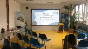 Ein großer, freundlich gestalteter und lichtdurchfluteter Seminarraum mit großer Präsentationsfläche und locker verteilten, blau gepolsterten Stühlen.