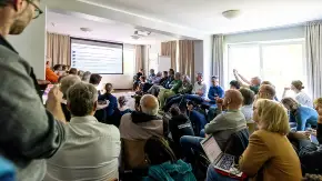 In einen Seminarraum schauen zahlreiche Zuschauende interessiert auf die Bühne, wo gerade eine Präsentation vorgeführt wird.