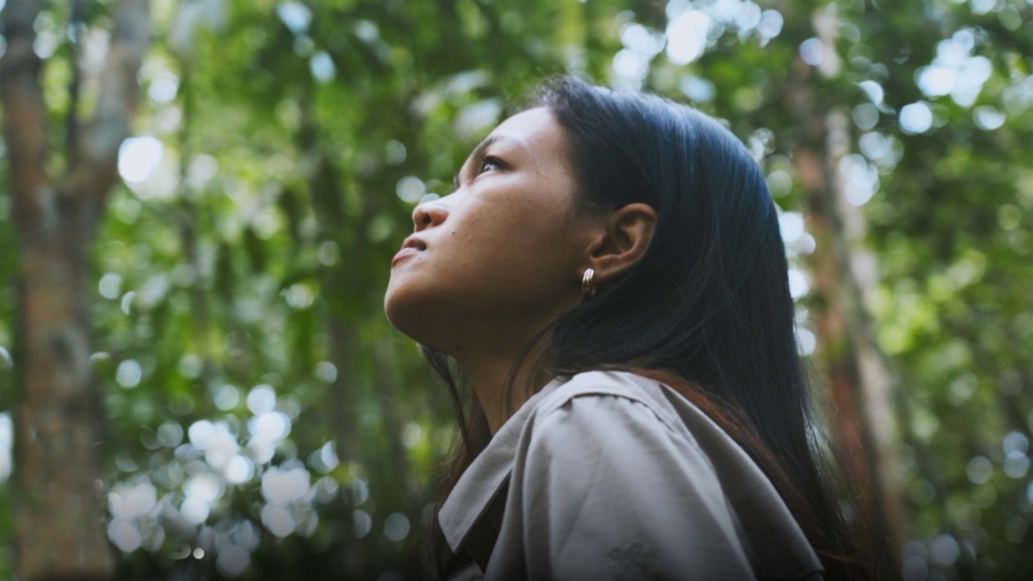 Eine junge Frau indigener Herkunft steht in einem Wald und schaut nach links oben.