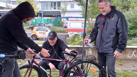 Drei Männer stehen in einem Park um ein Fahrrad herum, das sie begutachten.