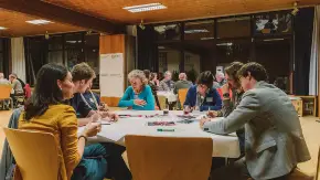 Ein großer Seminarraum mit holzverkleideter Decke, an Tischen sitzen Menschen unterschiedlichen Alters in Arbeitsgruppen