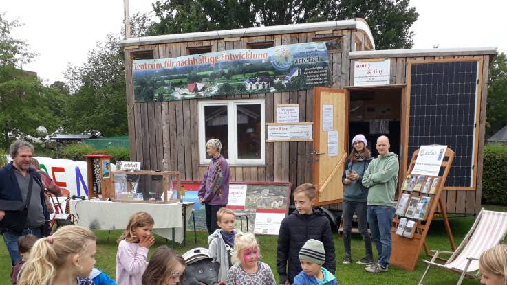 Ein kleines Holzhaus mit Fensern und einem Bsnner auf einem Anhänger auf einer Veranstaltung, davor ein Aufsteller mit Infomaterialien und Menschen und eine Gruppe Kinder 