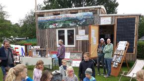 Ein kleines Holzhaus mit Fensern und einem Bsnner auf einem Anhänger auf einer Veranstaltung, davor ein Aufsteller mit Infomaterialien und Menschen und eine Gruppe Kinder 