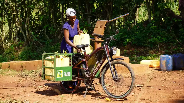 Eine Schwarze Frau mit einem beladenen E-Bike der Marke AfricroozE vor Bäumen