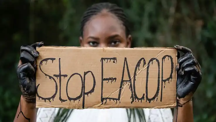 Eine Schwarze Frau mit ölverschmierten Händen hält einen Karton, auf dem «Stop EACOP» steht.