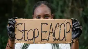 Eine Schwarze Frau mit ölverschmierten Händen hält einen Karton, auf dem «Stop EACOP» steht.