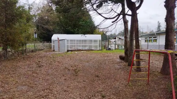 Ein Gewächshaus, links davon Bäume, rechts ein einstöckiges Gebäude. Im Vordergrund weitere Bäume, darunter ein rotgelbes Klettergerüst für Kinder.