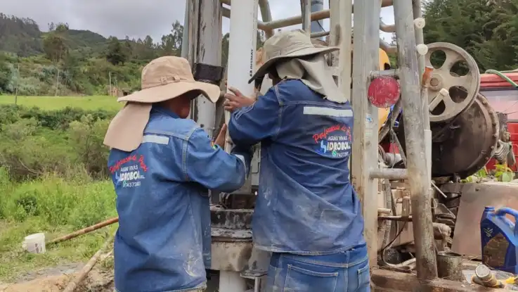 Zwei Menschen mit Kopfbedeckungen und Nackenschutz hantieren mit Bohrgestänge, auf ihren Arbeitskitteln ist der Name einer Baufirma abgebildet