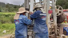 Zwei Menschen mit Kopfbedeckungen und Nackenschutz hantieren mit Bohrgestänge, auf ihren Arbeitskitteln ist der Name einer Baufirma abgebildet