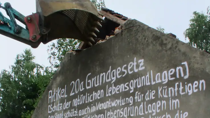 Ein Bagger ist im Begriff , eine kleine Hausruine abzureißen, auf dessen Giebelseite jemand groß den Grundgesetzartikel zum Schutz der natürlichen Lebensgrundlagen gepinselt hat.