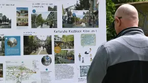Ein glatzköpfiger Mann mit Sonnenbrille steht im Freien an einer Straße und studiert, von der fotografierenden Person abgewandt, eine Wandzeitung zum Thema Kiezblocks.