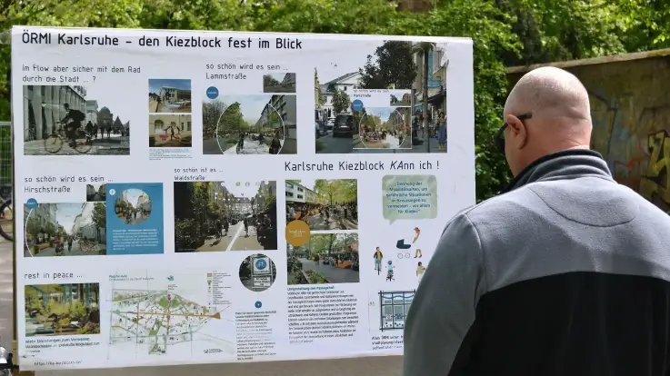 Ein glatzköpfiger Mann mit Sonnenbrille steht im Freien an einer Straße und studiert, von der fotografierenden Person abgewandt, eine Wandzeitung zum Thema Kiezblocks.
