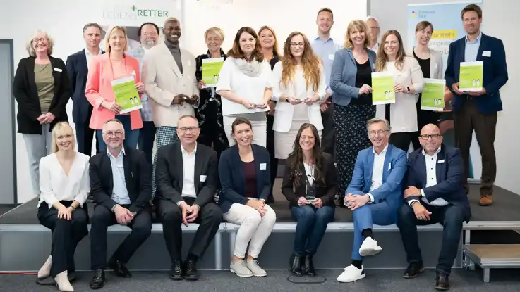 Eine große Gruppe Menschen posiert teils stehend, teils sitzend für ein Foto, fünf Personen darunter halten eine Auszeichnung in der Hand.