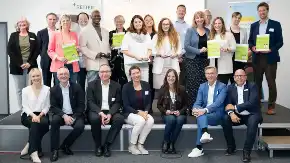 Eine große Gruppe Menschen posiert teils stehend, teils sitzend für ein Foto, fünf Personen darunter halten eine Auszeichnung in der Hand.