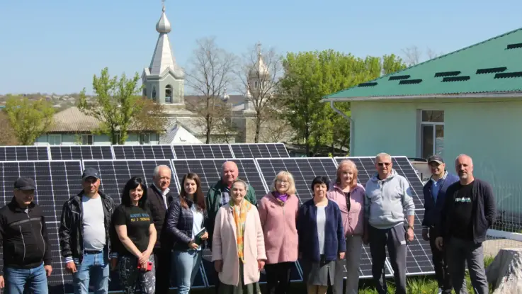 Eine Menschengruppe steht vor PV-Modulen, im Hintergrund sind Gebäude mit Zwiebeltürmen und die Shilouette einer Stadt zu erkennnen.