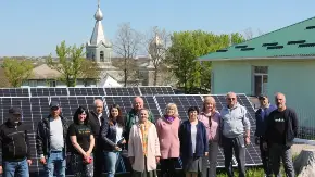 Eine Menschengruppe steht vor PV-Modulen, im Hintergrund sind Gebäude mit Zwiebeltürmen und die Shilouette einer Stadt zu erkennnen.