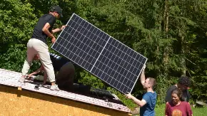 Ein Junger Mann reicht einem anderen Jungen Mann, der auf dem Dach eines Schuppen steht, ein ein PV-Modul zu.