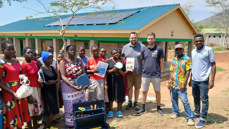 Vor einem eingeschossigen Gebäude mit PV-Modulen auf dem Dach steht eine Gruppe Menschen, teils afrikanischer, teils europäischer Herkunft. Einige halten Gegenstände wie eine Kochplatte oder ein Mini-PV-Modul in der Händen.