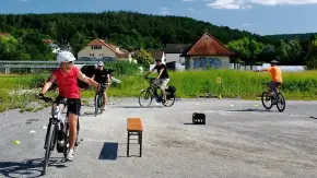 Auf einer geschotterten Fläche ist ein Parcour abgesteckt; drei Menschen auf E-Bike fahren hinter einem vierten her, der sich zur Seite dreht und etwas erläutert. Im Hintergrund ein kleiner Ort und waldige Hügel unter blauem Himmel.