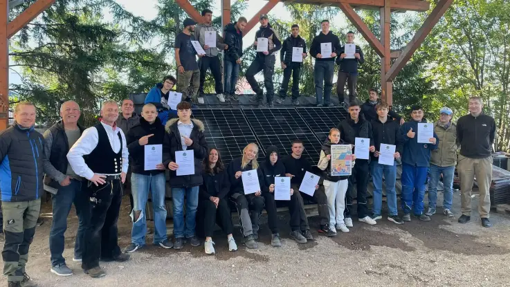 Rund zwei Dutzend Teilnehmende un d Lehrpersonen haben sich vor, neben und auf eine schräggestellten PV-Modulfläche für ein Gruppenfoto aufgestehlz