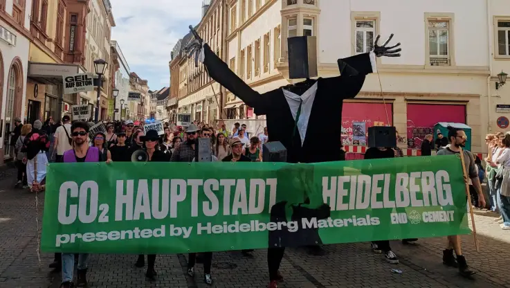 Protestzug durch die Innenstadt, vorne eine Puppe mit Lautsprecherkopf und ein großes grünes Banner mit dem weiß gehaltenen Slogan «CO2-Hauptstadt Heidelberg»