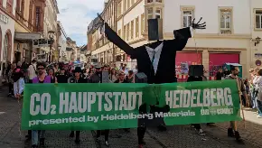 Protestzug durch die Innenstadt, vorne eine Puppe mit Lautsprecherkopf und ein großes grünes Banner mit dem weiß gehaltenen Slogan «CO2-Hauptstadt Heidelberg»