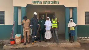 Vor einem eingeschossigen Gebäude mit PV-Modulen auf dem Dach steht eine Gruppe Menschen, teils afrikanischer, teils europäischer Herkunft. Einige halten Gegenstände wie eine Kochplatte oder ein Mini-PV-Modul in der Händen.