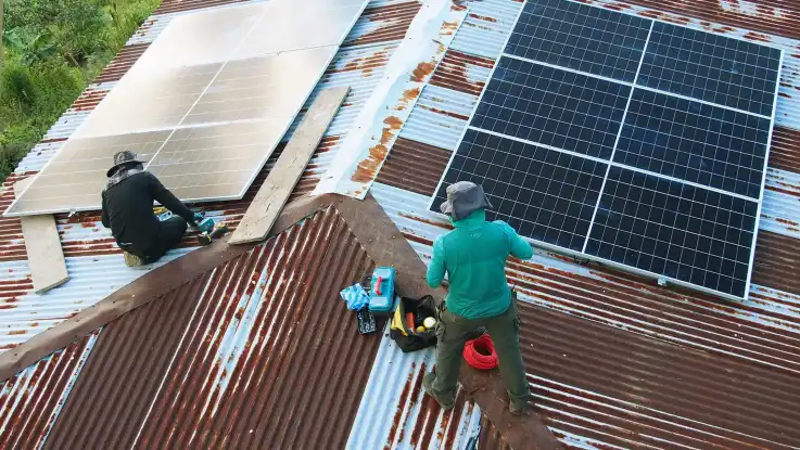 Zwei Techniker montieren auf dem Wellcllblechdach eines von tropisch anmutendem Wald umgebenen Hauses PV-Module.