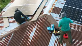 Zwei Techniker montieren auf dem Wellcllblechdach eines von tropisch anmutendem Wald umgebenen Hauses PV-Module.