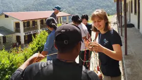 Oberhalb eines Schulgebäudes in bergiger Landschaft kämpfen unter einem Vordach zwei Personen unterschiedlicher Herkunft lachend mit einem Kabelgewirr.