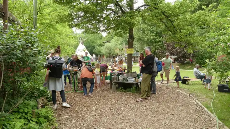 Kinder und Erwachsene in einem parkartigen Grünbereich, ein Mann strampelt auf einen Energiefahrrad, andere schauen zu. Im HIntergrund ein Tipi.