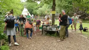 Kinder und Erwachsene in einem parkartigen Grünbereich, ein Mann strampelt auf einen Energiefahrrad, andere schauen zu. Im HIntergrund ein Tipi.
