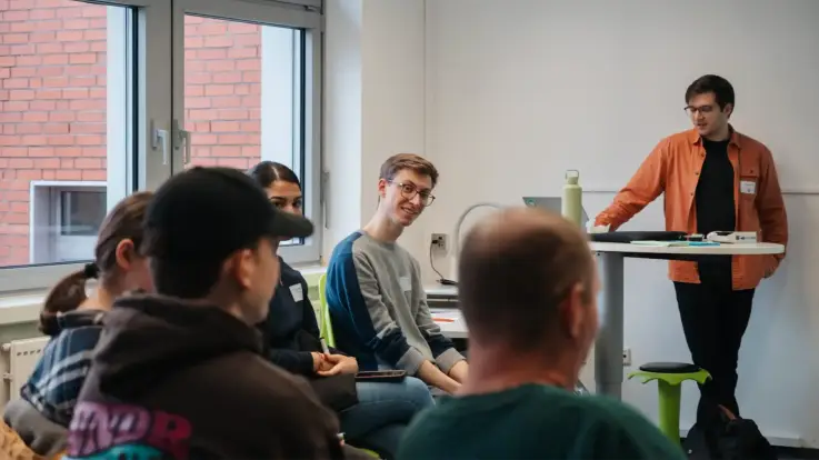 In einem Schulraum referiert ein junger Mann mit orangefarbenem Hemd, an ein Pult gelehn, vor einigen Schüler:innen und einer Lehrperson.
