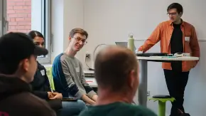 In einem Schulraum referiert ein junger Mann mit orangefarbenem Hemd, an ein Pult gelehn, vor einigen Schüler:innen und einer Lehrperson.