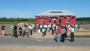 Eine Menschengruppe steht vor einem roten Haus, das einzeln in einer Ebene steht
