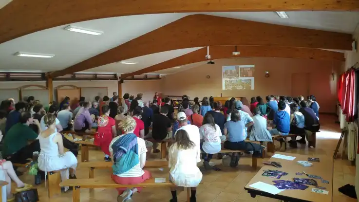 In einem großen Seminarraum mit einer Deckenkonstruktion aus Holz sitzen zahlreiche Menschen auf Bänken und verfolgen eine Präsentation.