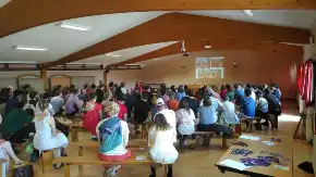 In einem großen Seminarraum mit einer Deckenkonstruktion aus Holz sitzen zahlreiche Menschen auf Bänken und verfolgen eine Präsentation.