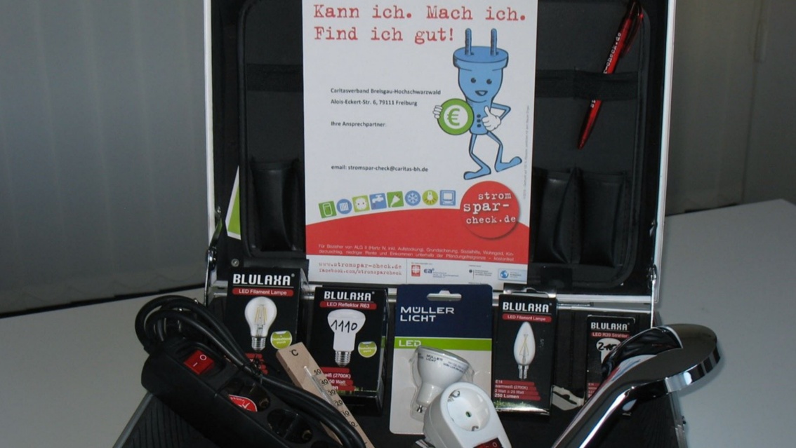 Ein gepolsterter Aluminiumkoffer, im aufgeklappten Teil ein Plakat mit Werbung für den Stromspar-Check, darin Hilfsmittel zum Energiesparen wie abschaltbare Steckdosen, LED-Lampen und ein wassersparender Duschkopf