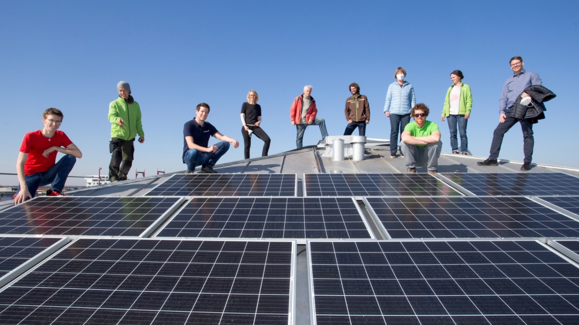 Eine Gruppe von zehn Menschen steht bei strahlendem Sonnenschein auf einem leicht geneigten Blechdach, im Vordergrund zahlreiche PV-Module.