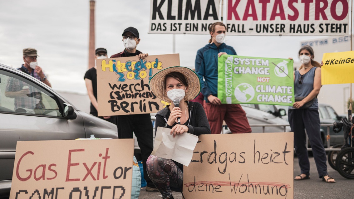 Eine Gruppe junger Aktivist:innen posiert mit Plakaten gegen die Klimapolitik zwischen parkenden Autos; im Vordergrund kauert eine junge Frau mit Strohhut, Gesichtsmaske und Mikrofon und präsentiert zwei weitere handgeschriebene Plakate.