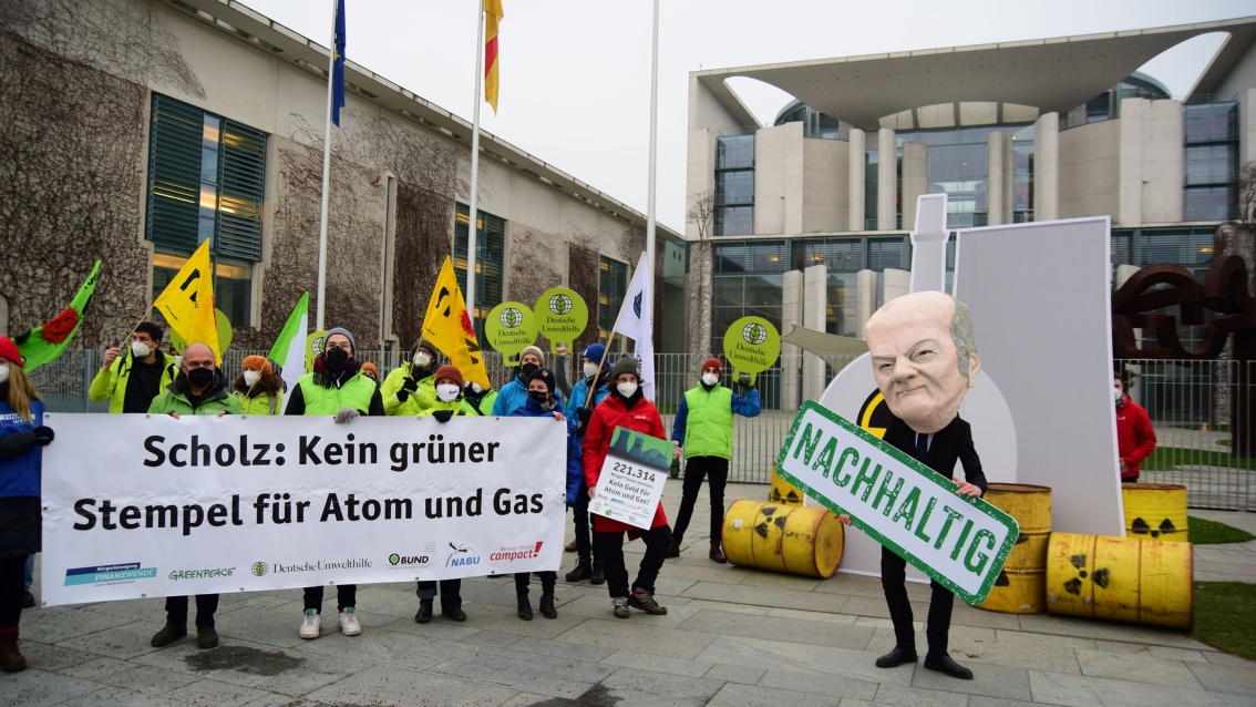 Vor dem Kanzleramt in Berlin präsentieren Aktivistinnen ein Banner, das Olaf Scholz auffordert, Atomkraft und Gas keinen grünen Stempel zu verleihen; rechts im Bild ein einzeln stehender Mensch mit übergroßer Olaf-Scholz-Maske, der ein «Nachhaltig»-Schild in der Hand hält.