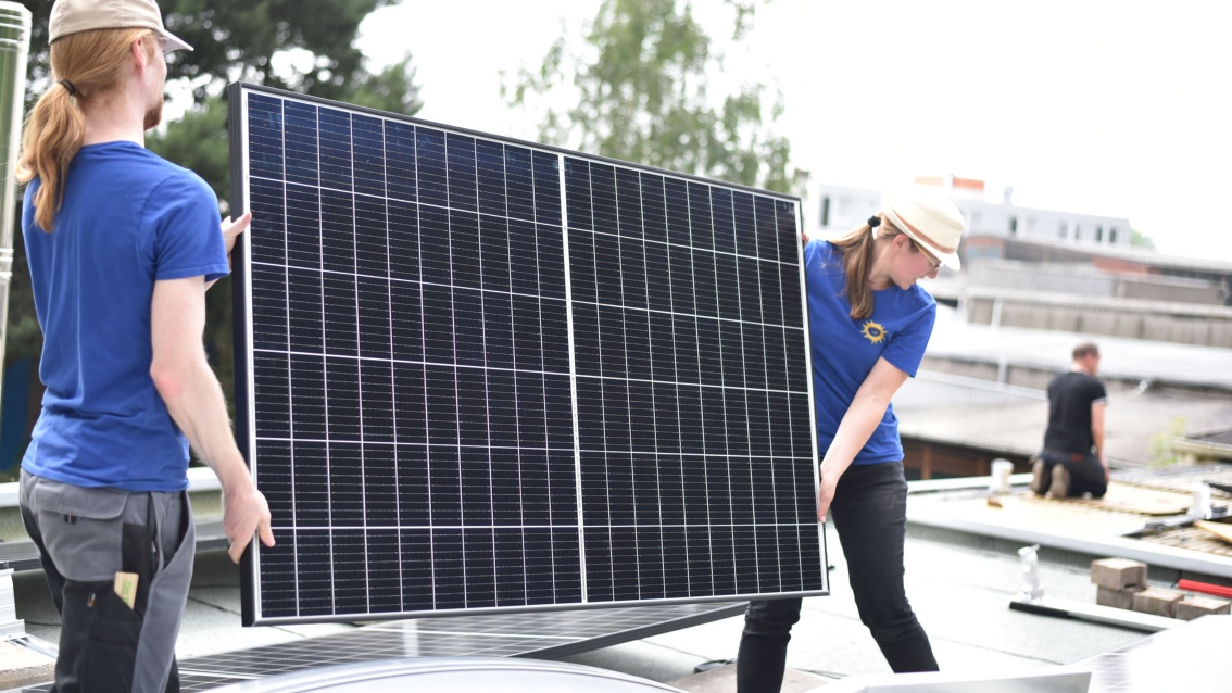 Zwei junge Menschen auf einem Hausdach tragen behutsam ein großes Photovoltaikmodul in Richtung eines im Hintergrund knienden Menschen.