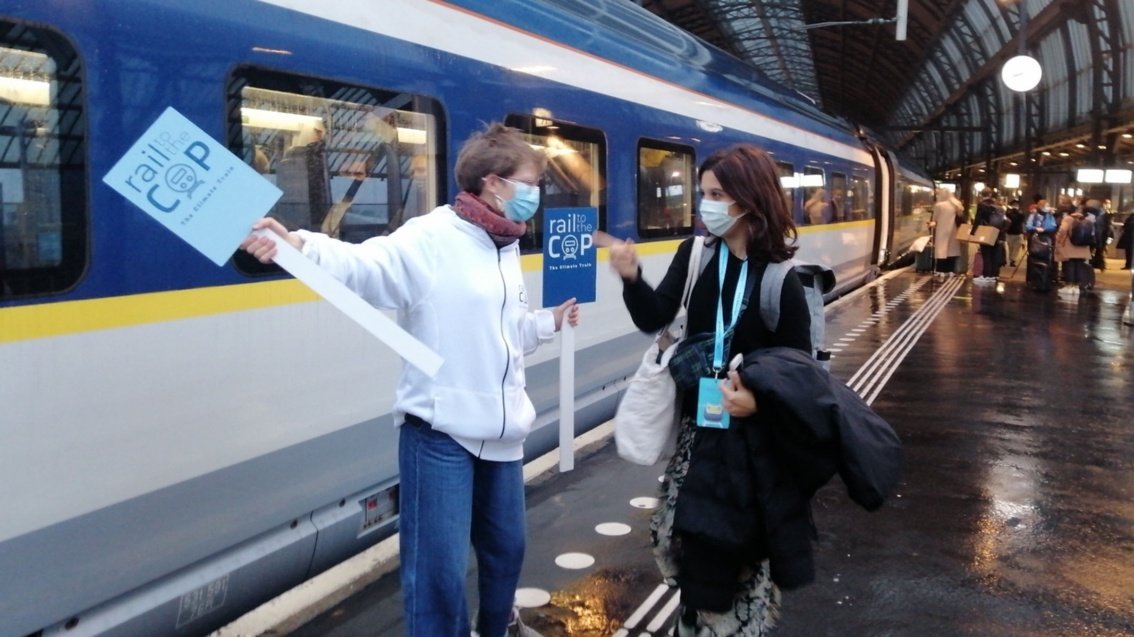 An einem regennassen Bahnsteig vor einem bereitstehenden Schnellzug weist eine Frau mit einem Schild «Rail to the COP» einer anderen Frau den Weg.