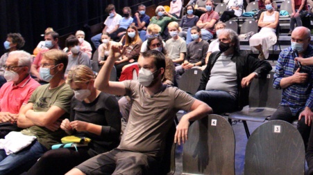 In einer großen Veranstaltungshalle sitzen Menschen mit Gesichtsmasken auf den nach hinten ansteigenden Sitzreihen und blicken nach links vorne in Richtung der Bühne; ein junger, leger gekleideter Mann in der ersten Reihe medet sich zu Wort.