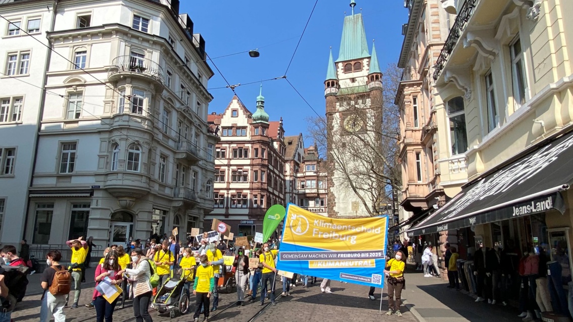 Auf einer der Straßen, die vom wuchtigen Freiburger Martinstor mit seinem von fünf Zinnen gekrönten Turm wegführen, wirbt eine große Gruppe vorwiegend gelb gekleideter Menschen mit einem großen Banner für einen Klimaentscheid in der Breisgaumetropole.