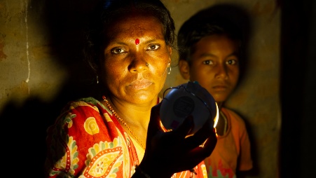 Eine indische Frau hält eine eine kleine Solarleuchte so vor sich, dass diese ihr Gesicht sowie das eines im Hintergrund stehenden Kindes erhellt.